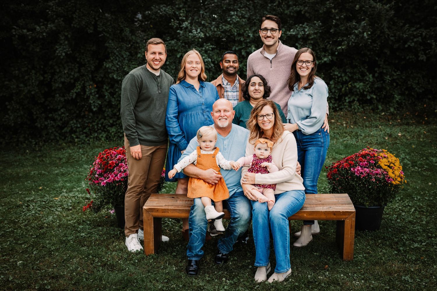 Group sitting together on wooden bench outdoors with colorful flowers in background during autumn photo session.