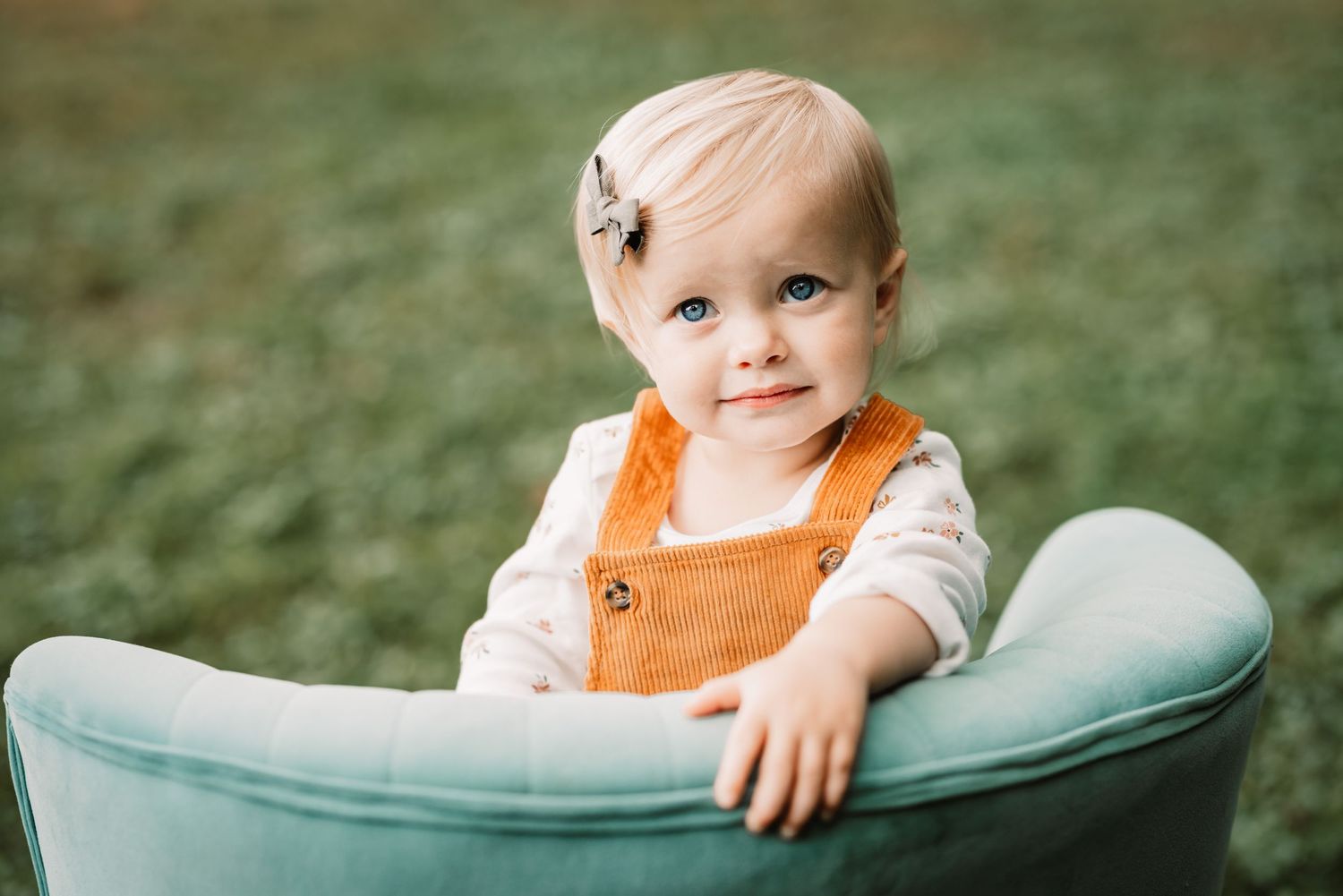 Baby wearing orange overalls sits in mint green chair outdoors.