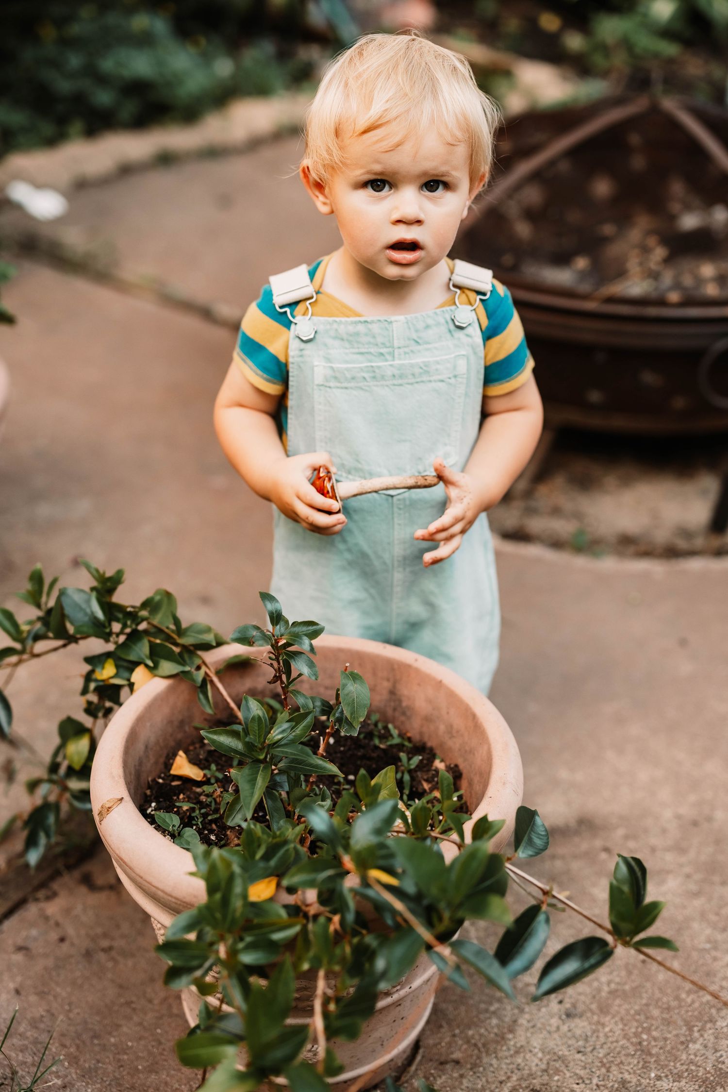 Small child in aqua dress explores potted plants while sitting on a concrete surface outdoors.