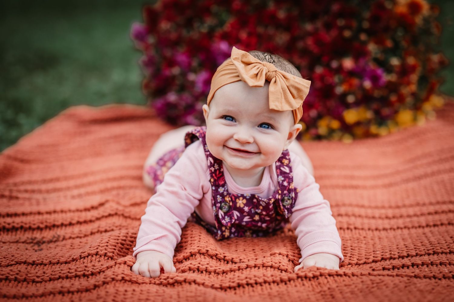 Baby wearing floral dress and bow headband smiles while laying on orange blanket outdoors.
