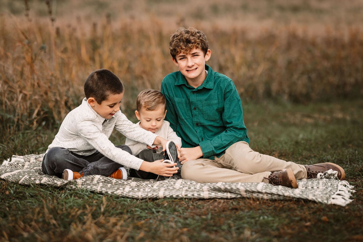 Three children sit on a blanket outdoors looking at something together.