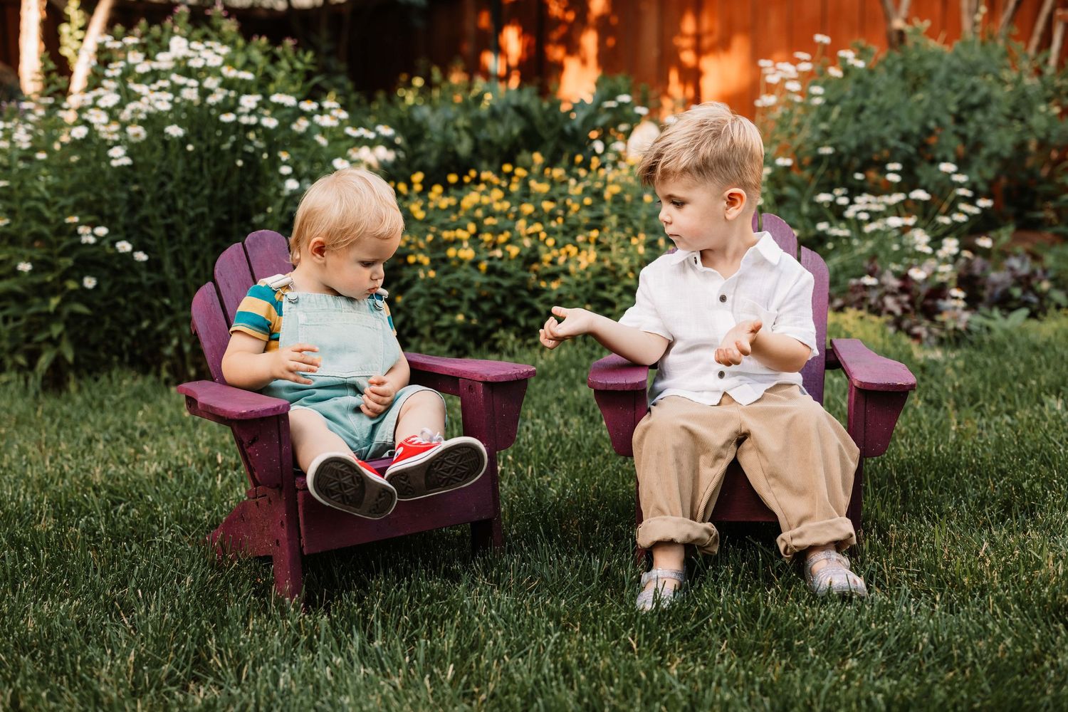 Two kids sit together reading books in small burgundy chairs on a grassy lawn with flowers in the background.
