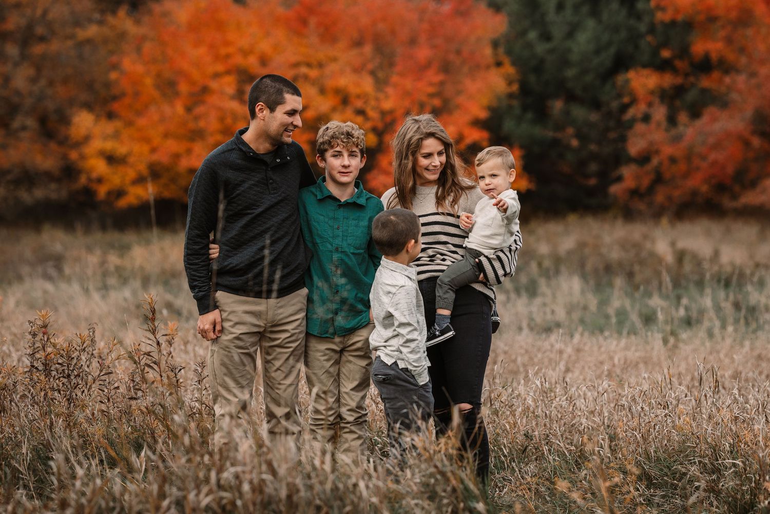 A family of four poses together in a field with bright orange autumn trees behind them.