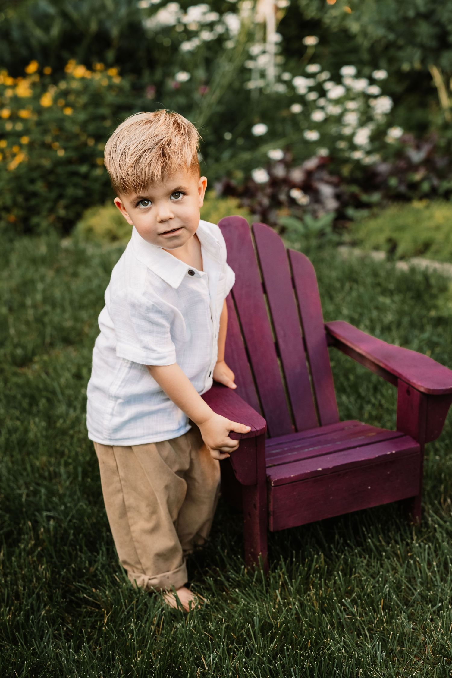 In a garden setting, a purple wooden Adirondack chair rests on green grass surrounded by summer flowers.