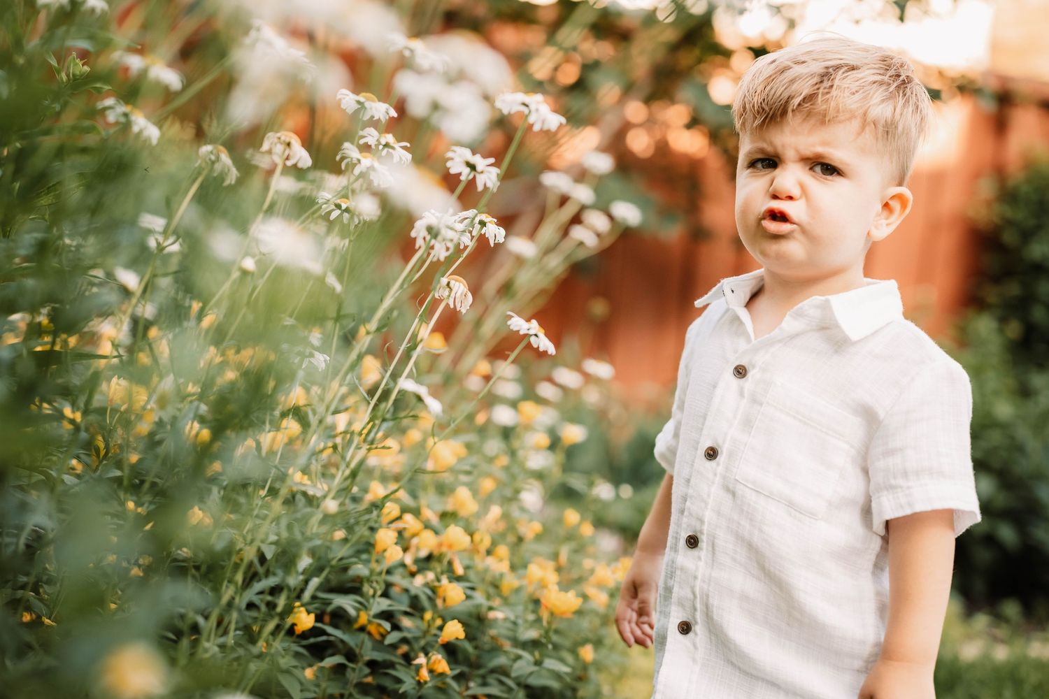Child in white linen shirt stands thoughtfully among blooming summer daisies in a garden setting.