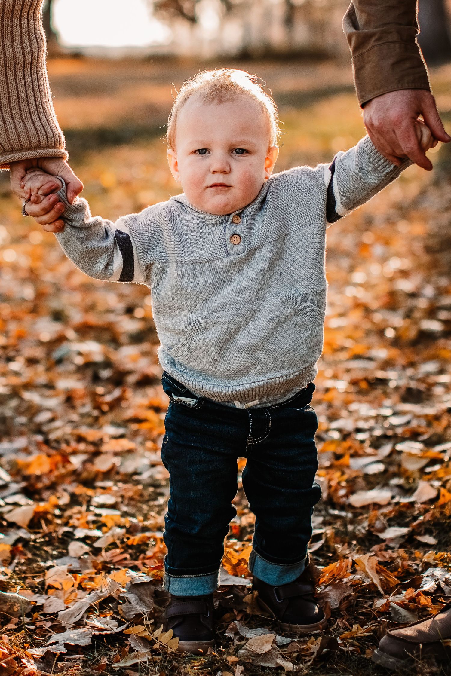 A small child in a grey sweater walks through fallen autumn leaves while holding hands with adults.