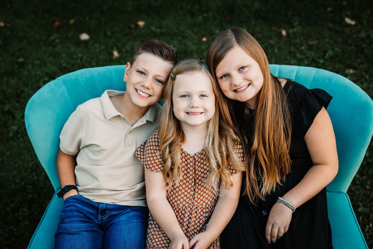 Three children sitting close together on turquoise vintage chair outdoors.
