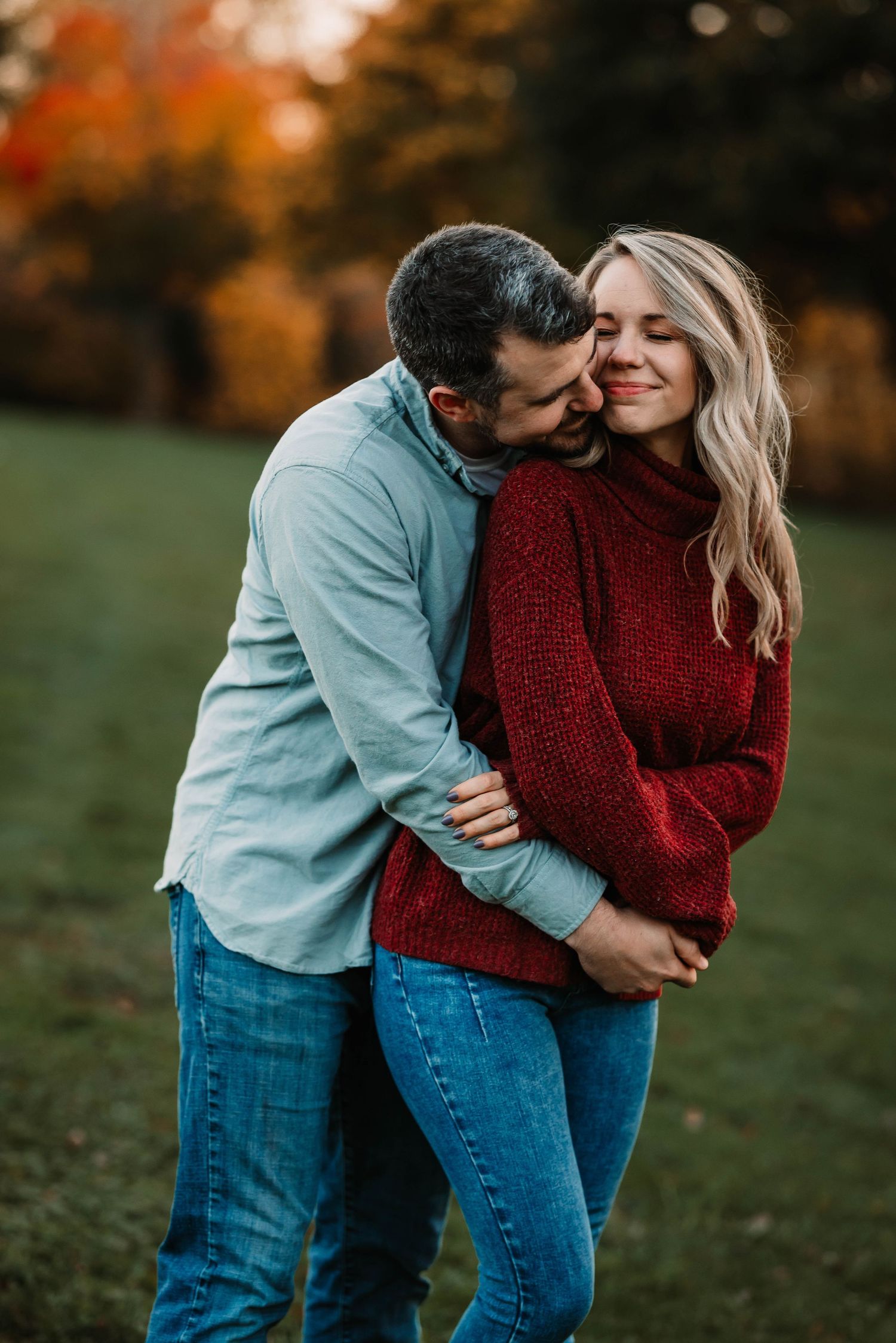 A couple in casual wear shares an intimate moment against autumn foliage.