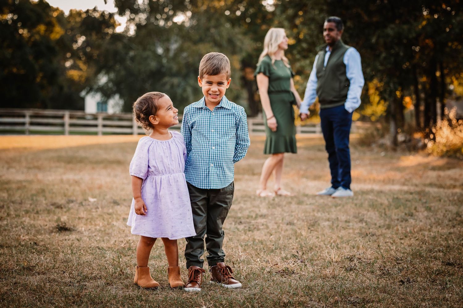 Two young children share a tender moment together in a field as adults stand in the background.