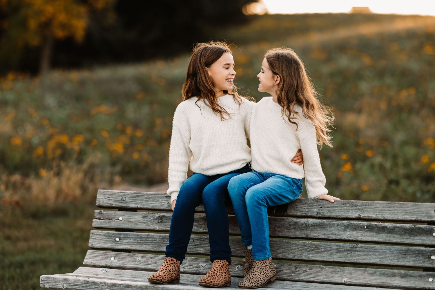 Two people in matching white sweaters and jeans sitting together on a wooden bench at sunset.