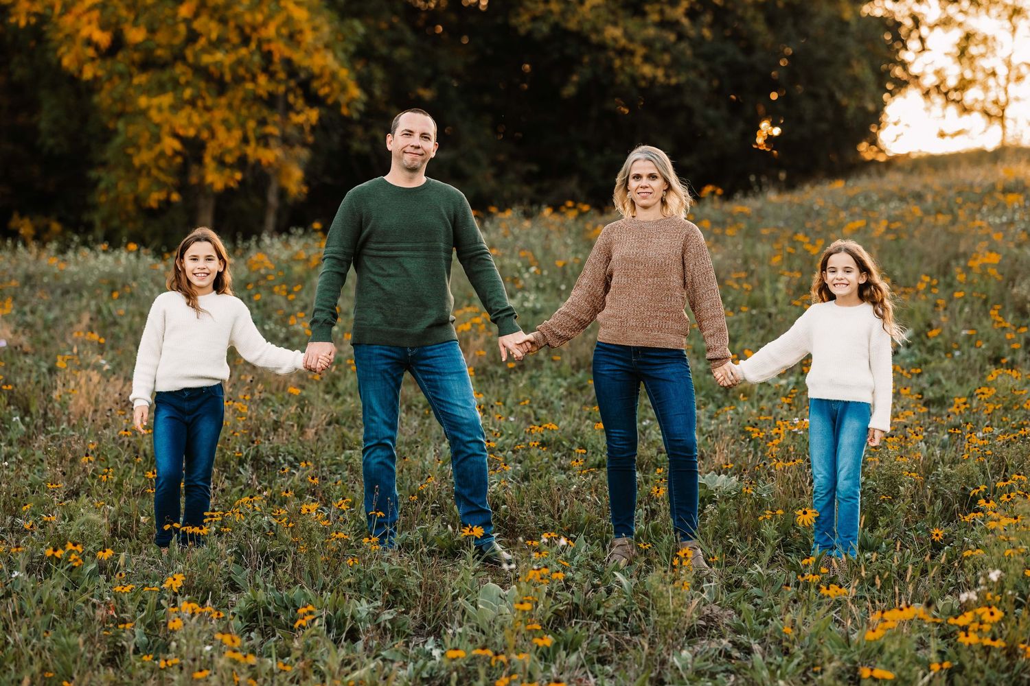Family of four holding hands while standing in a wildflower field at sunset.