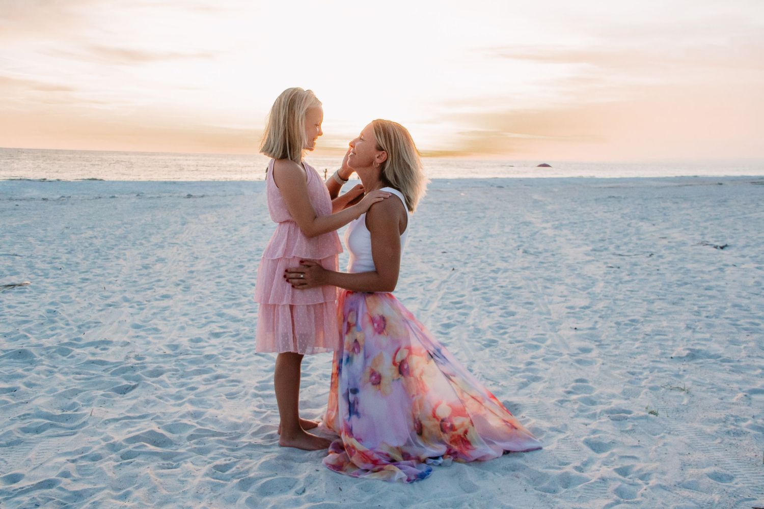 A tender moment between parent and child on a white sandy beach at sunset in matching pink dresses.