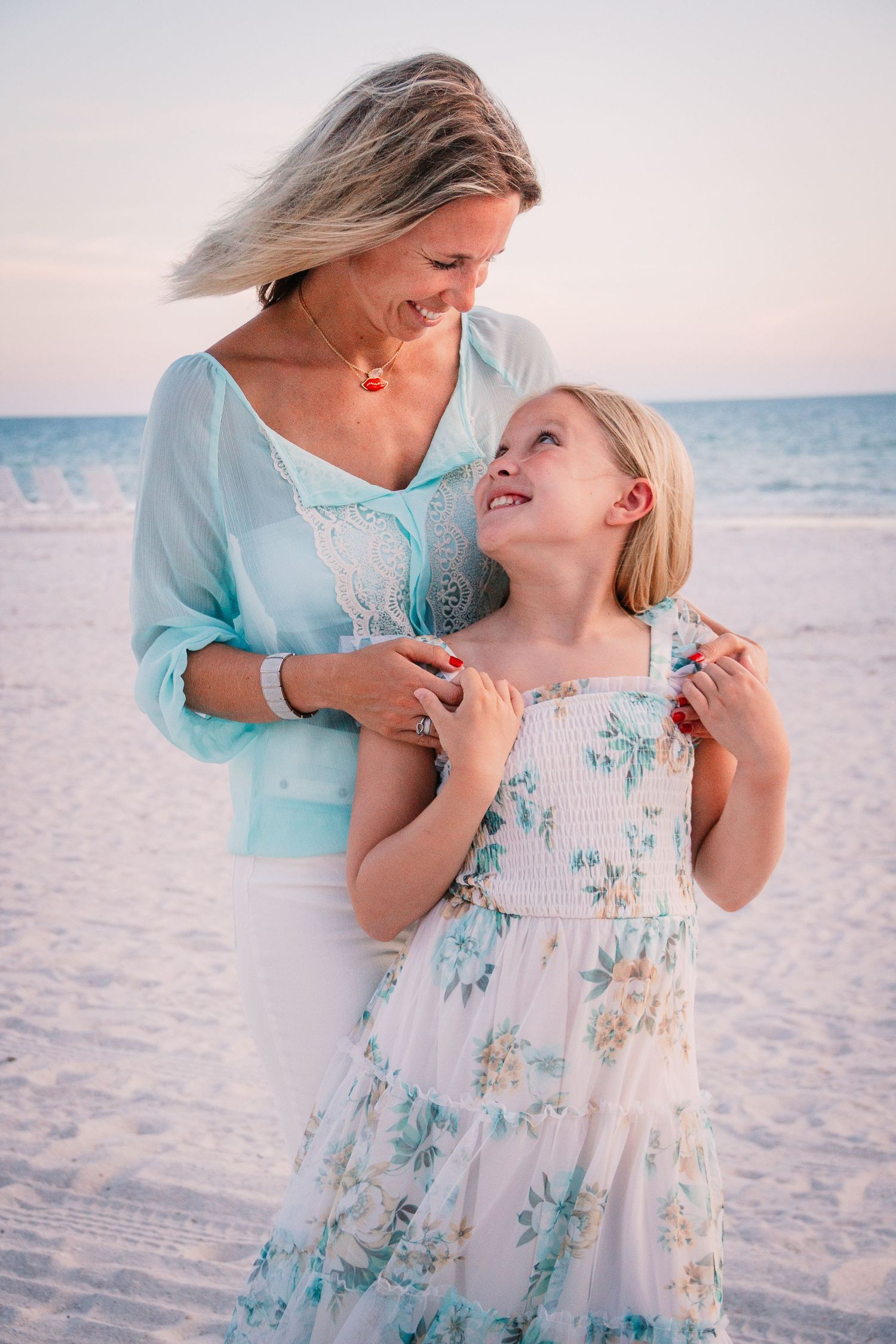 A tender beach moment is captured of two people in matching turquoise and white floral dresses against white sand at sunset.