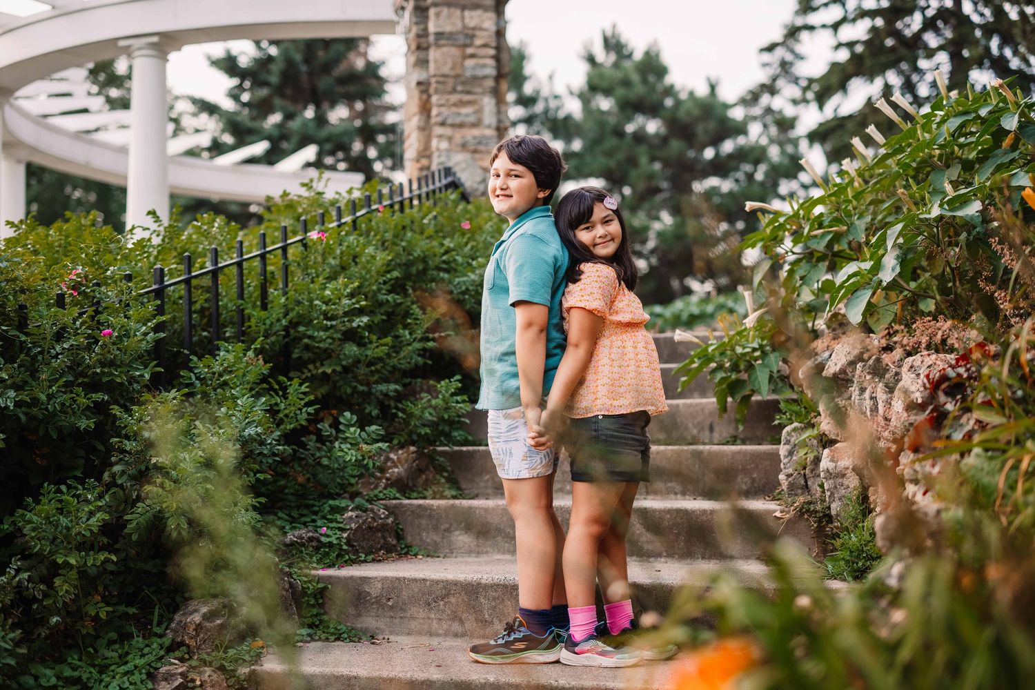 Two people stand together on stone steps with lush greenery and florals surrounding the outdoor scene.