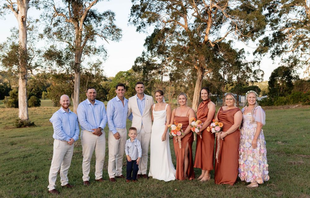 Wedding party posing outdoors on green lawn with eucalyptus trees, bride in white gown with bridesmaids in rust dresses.