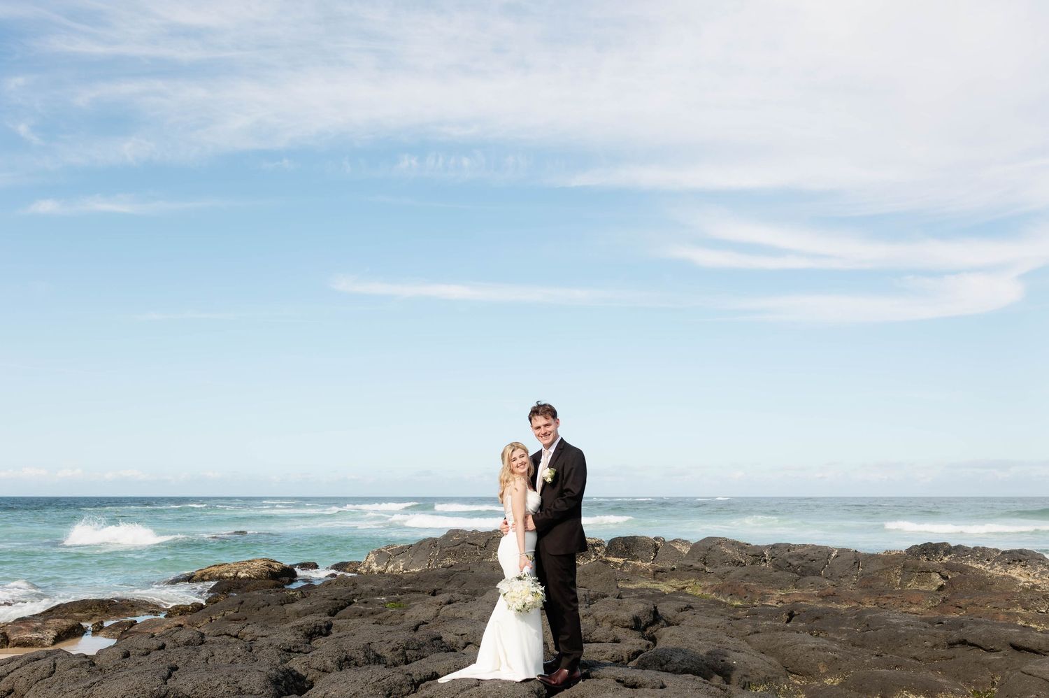 A couple in wedding attire embraces on rocky coastal cliffs with ocean waves and blue sky in the background.