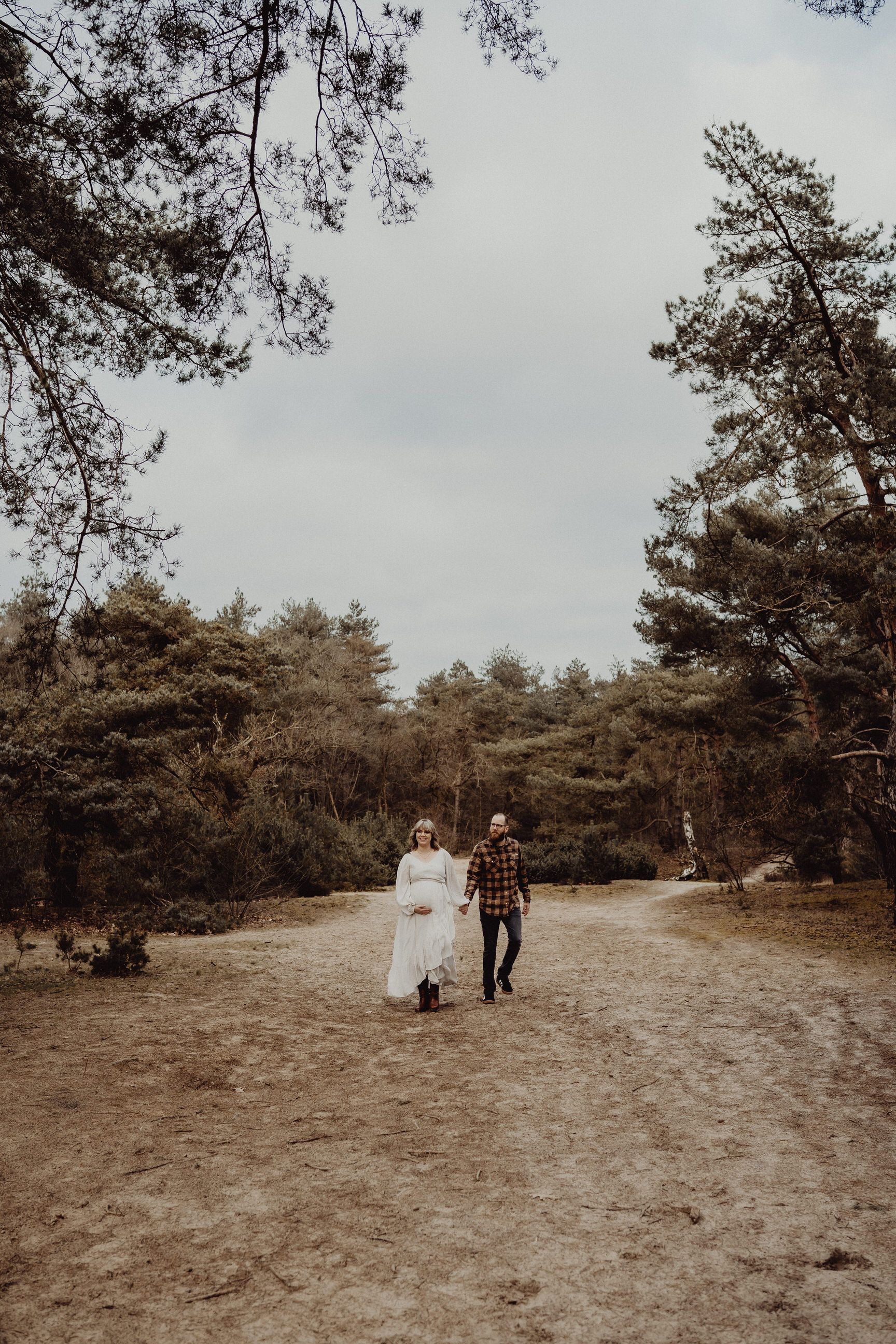 Two people walk down a sandy path surrounded by pine trees on a cloudy day.