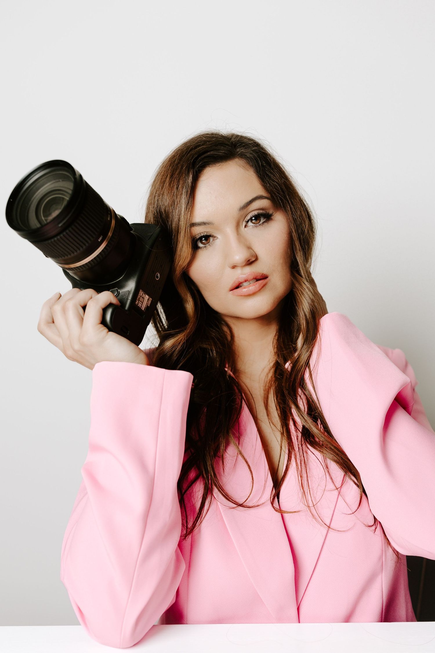 Professional photographer wearing pink blazer poses with camera against white background.