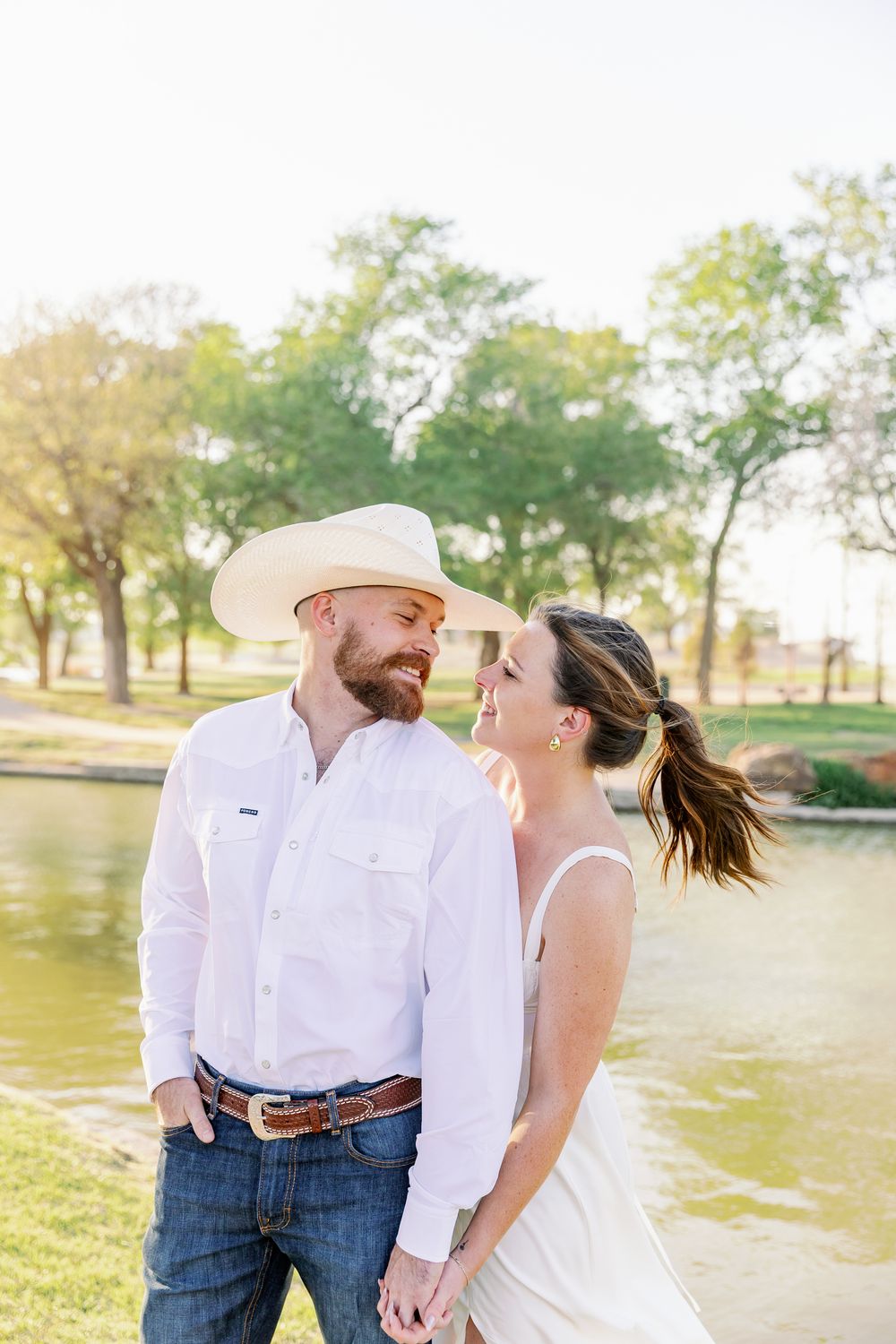 A couple in western attire embraces during a romantic sunset photoshoot in a grassy field near a lake.