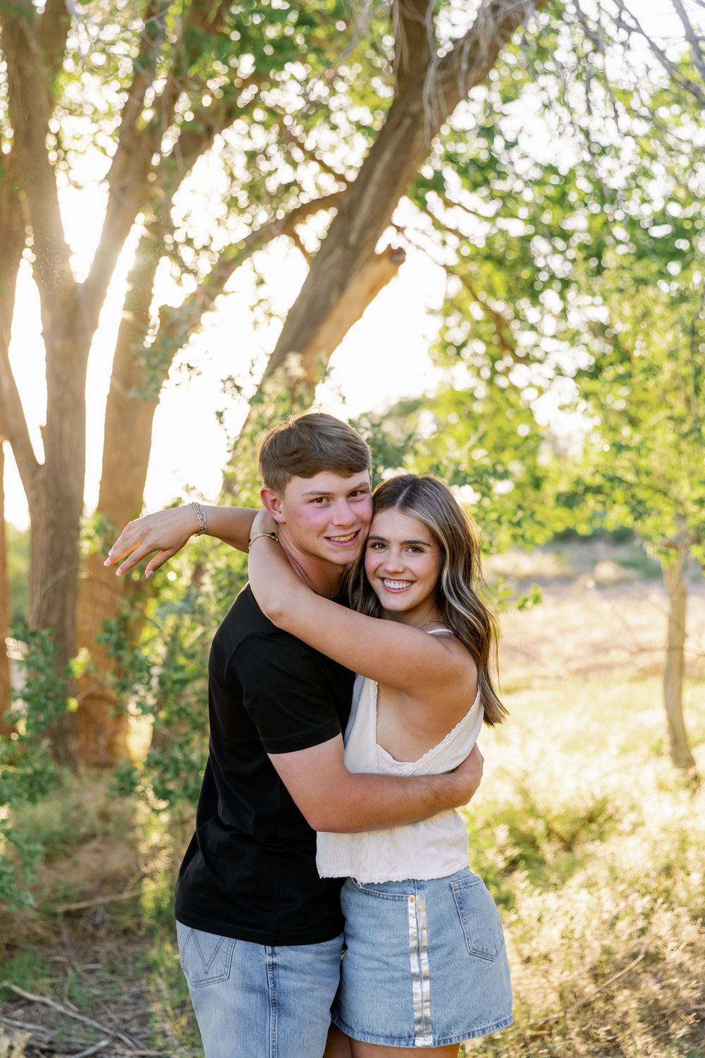 A couple embraces in a sunny wooded area, with sunlight filtering through trees in the background.