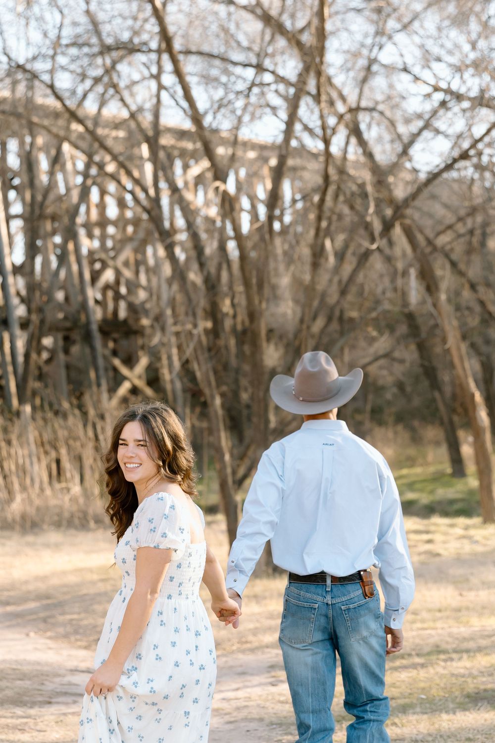 A couple holding hands walks together in a wooded area during sunset, person in white dress and person in denim.