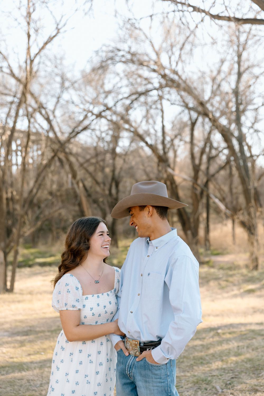A couple in western attire share playful moments while posing in a field with bare trees in the background.