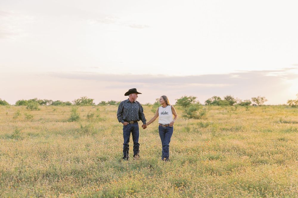 Two people walk through a golden wildflower meadow at sunset with rolling hills in the distance.