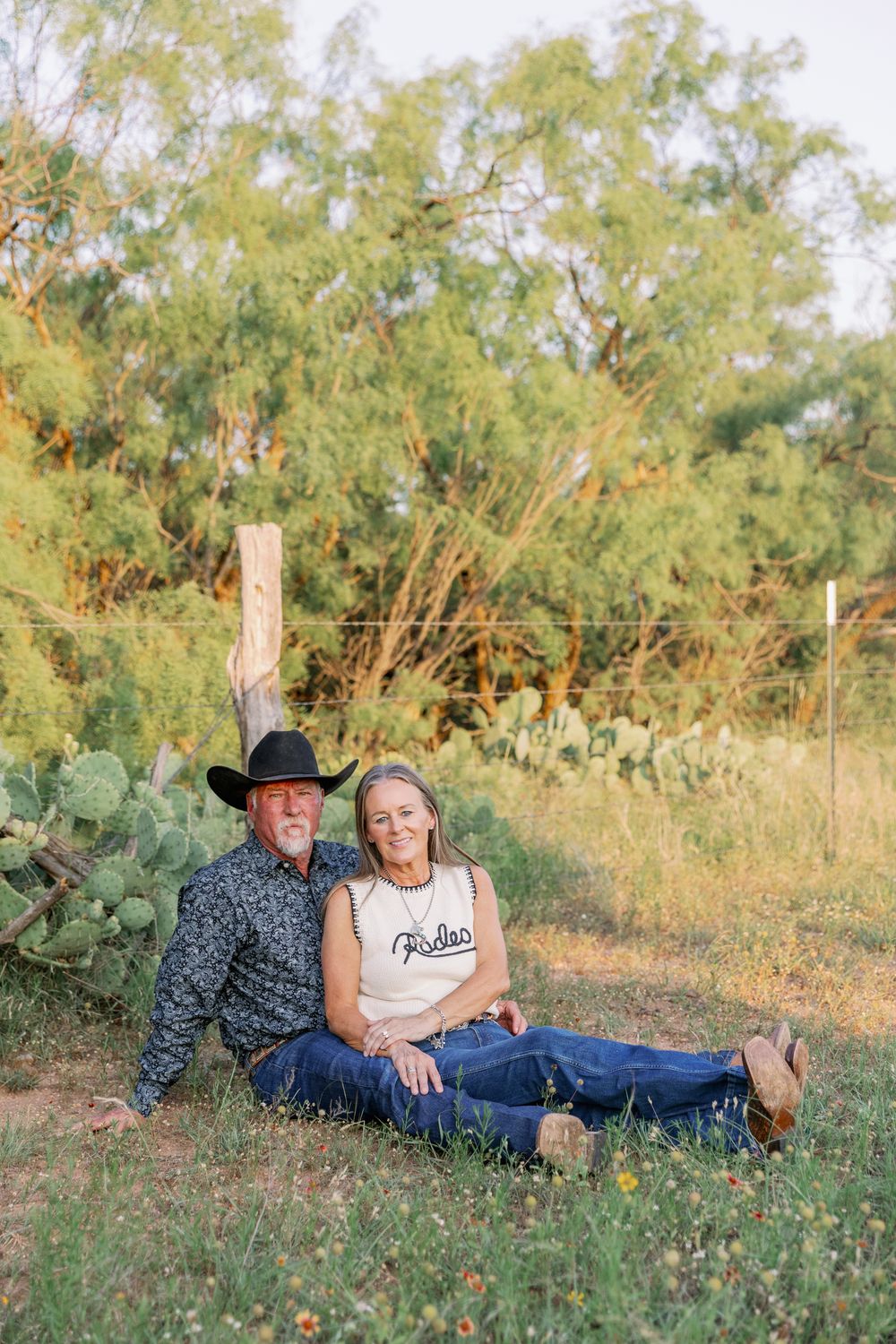 Two people relax together on grassy terrain with desert plants and trees in a casual outdoor setting.