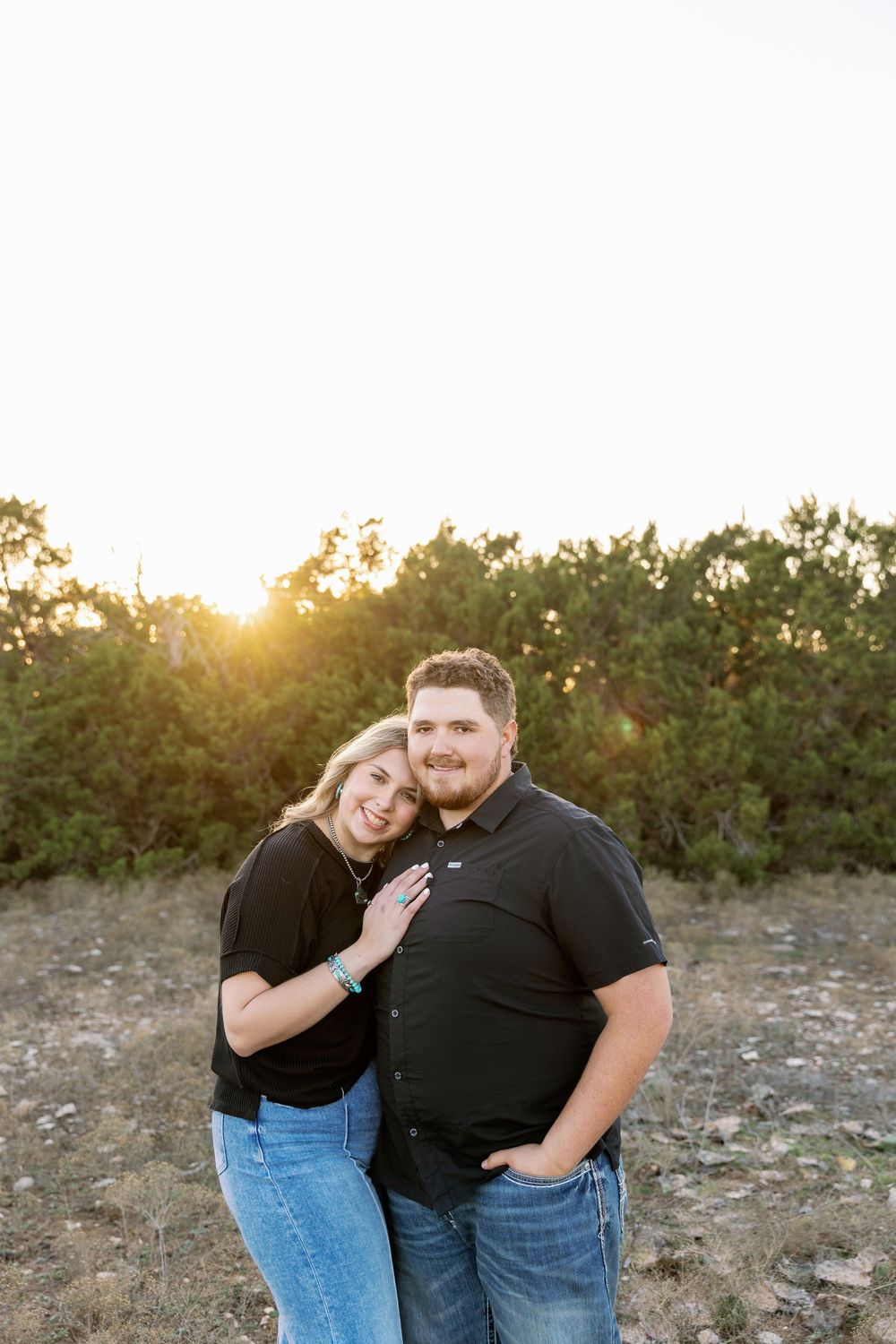 A couple embraces outdoors during golden hour sunset with trees in the background.