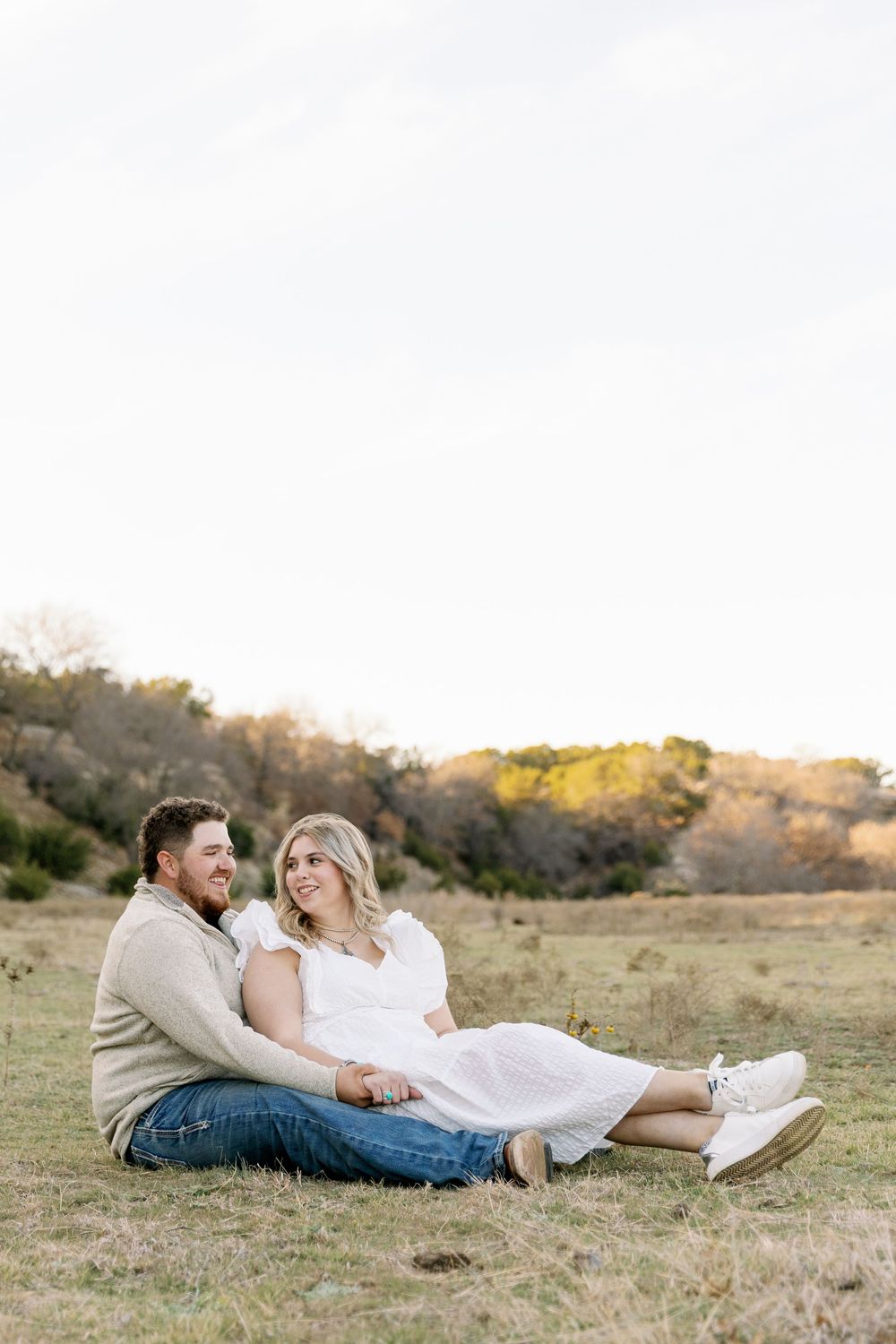 A couple relaxes together on a grassy hill during a romantic sunset photo session in an open field.