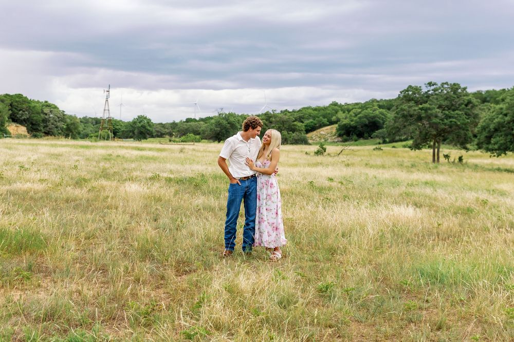 A couple embraces in a scenic meadow with rolling hills and trees under an overcast summer sky.