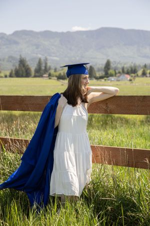 Cora Emilie Media Graduate in white dress and blue cap sits peacefully by wooden fence overlooking mountain landscape.