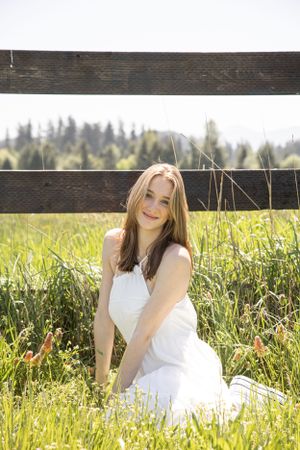 Cora Emilie Media Graduate in white dress reclines by wooden fence in sunny meadow with mountains behind.