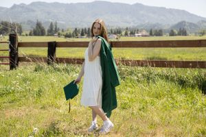 Cora Emilie Media Graduate in white dress and green gown walks through sunny meadow with mountains in background.
