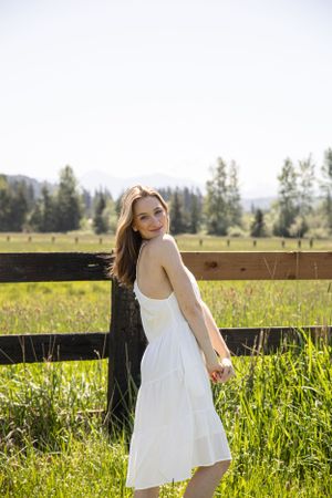 Cora Emilie Media Graduate in flowing white dress twirls joyfully in sunlit field by wooden fence.