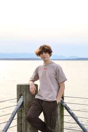 Cora Emilie Media Young person leaning on wooden pier post with water and mountains in background.