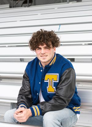 Cora Emilie Media Individual relaxes on metal bleachers wearing blue and gray varsity jacket with letter T.