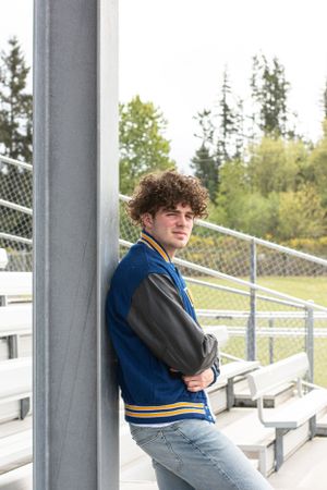 Cora Emilie Media Student in letterman jacket sits casually on stadium bleachers with trees in background.