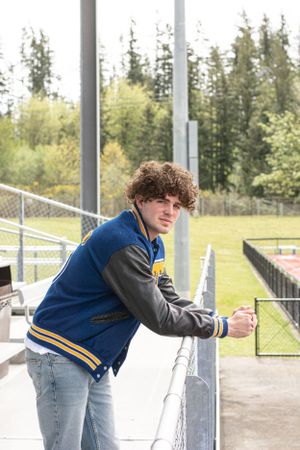 Cora Emilie Media Student in varsity letterman jacket leans against stadium bleachers surrounded by evergreen trees.