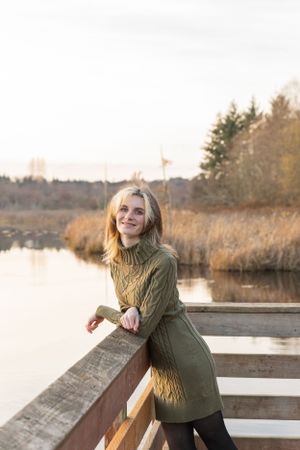 Cora Emilie Media Person in green sweater dress standing on wooden dock by lake at sunset.