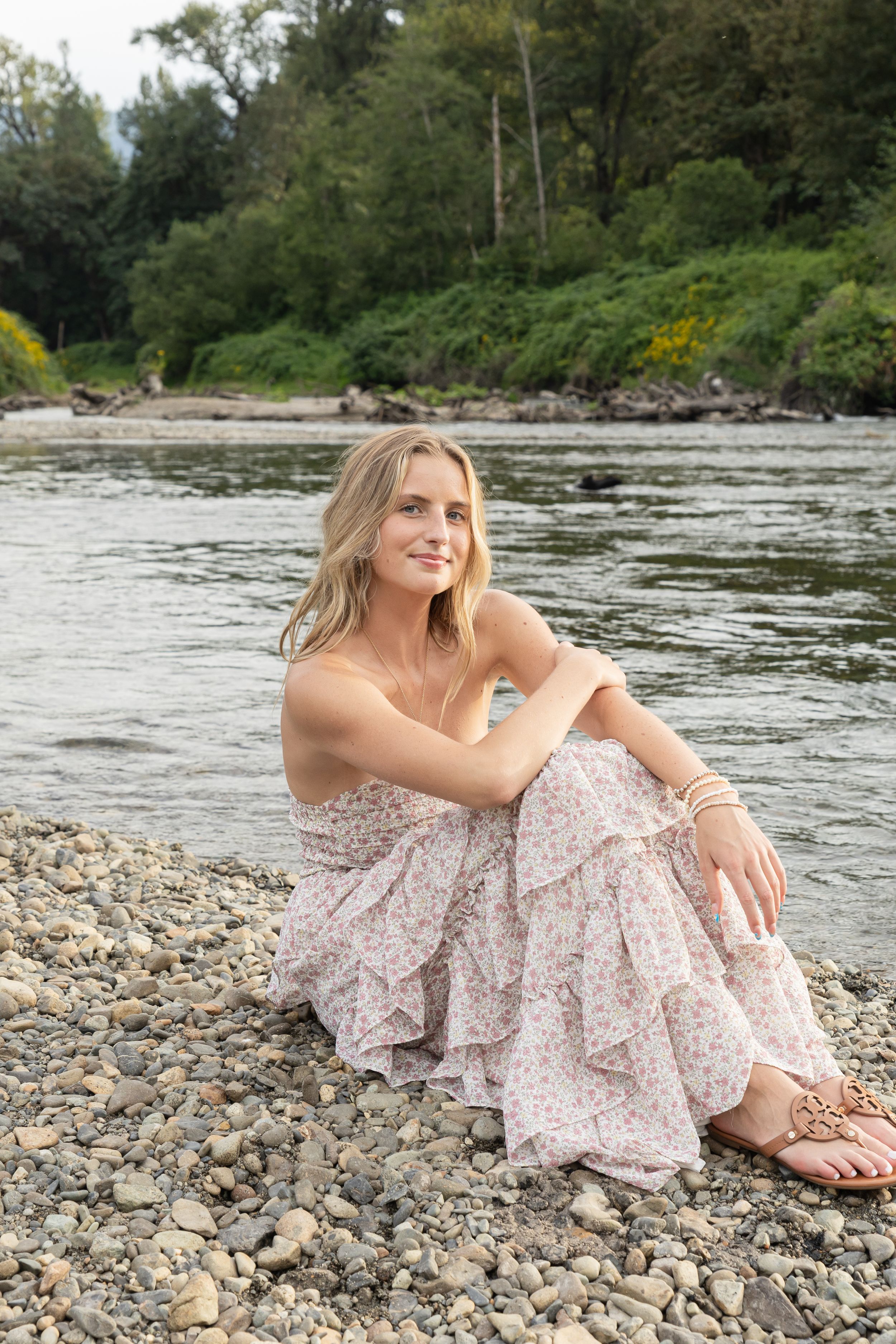 Cora Emilie Media Person in floral dress sitting on rocky riverbank with flowing water in background.