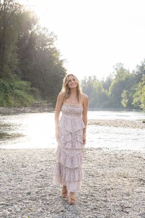 Cora Emilie Media Person in tiered floral maxi dress standing on rocky riverbank at sunset.