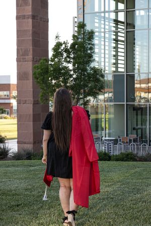 Cora Emilie Media Graduate in black dress walking on campus lawn with red graduation gown.