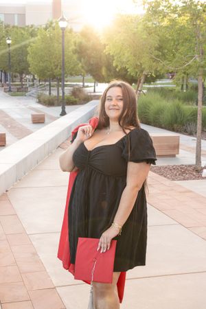 Cora Emilie Media Graduate in black dress holding red diploma folder on sunny campus walkway.