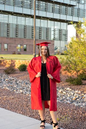 Cora Emilie Media Graduate posing in red cap and gown outside modern university building.