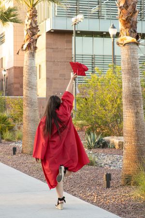 Cora Emilie Media Graduate in red cap and gown leaping joyfully between palm trees on campus.