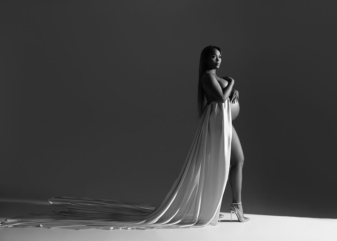 A dramatic black and white maternity photo showing a long flowing white dress trailing across a dark stage in Atlanta photography studio.