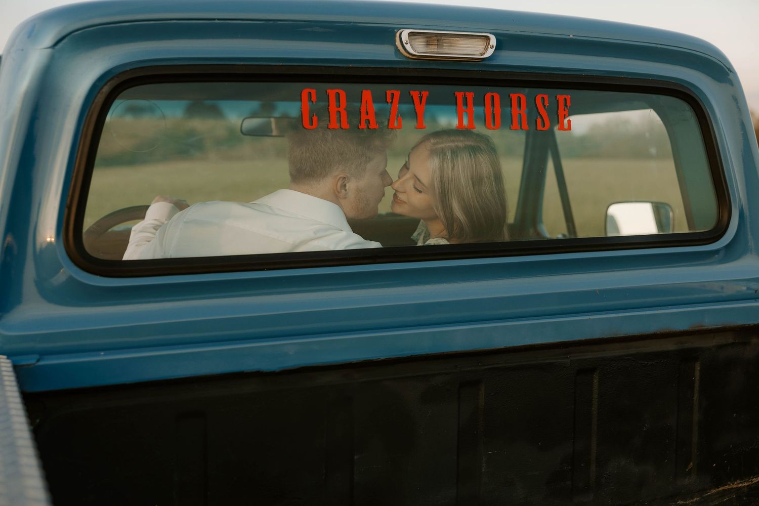 Couple posing beside vintage blue pickup truck in golden sunset field.