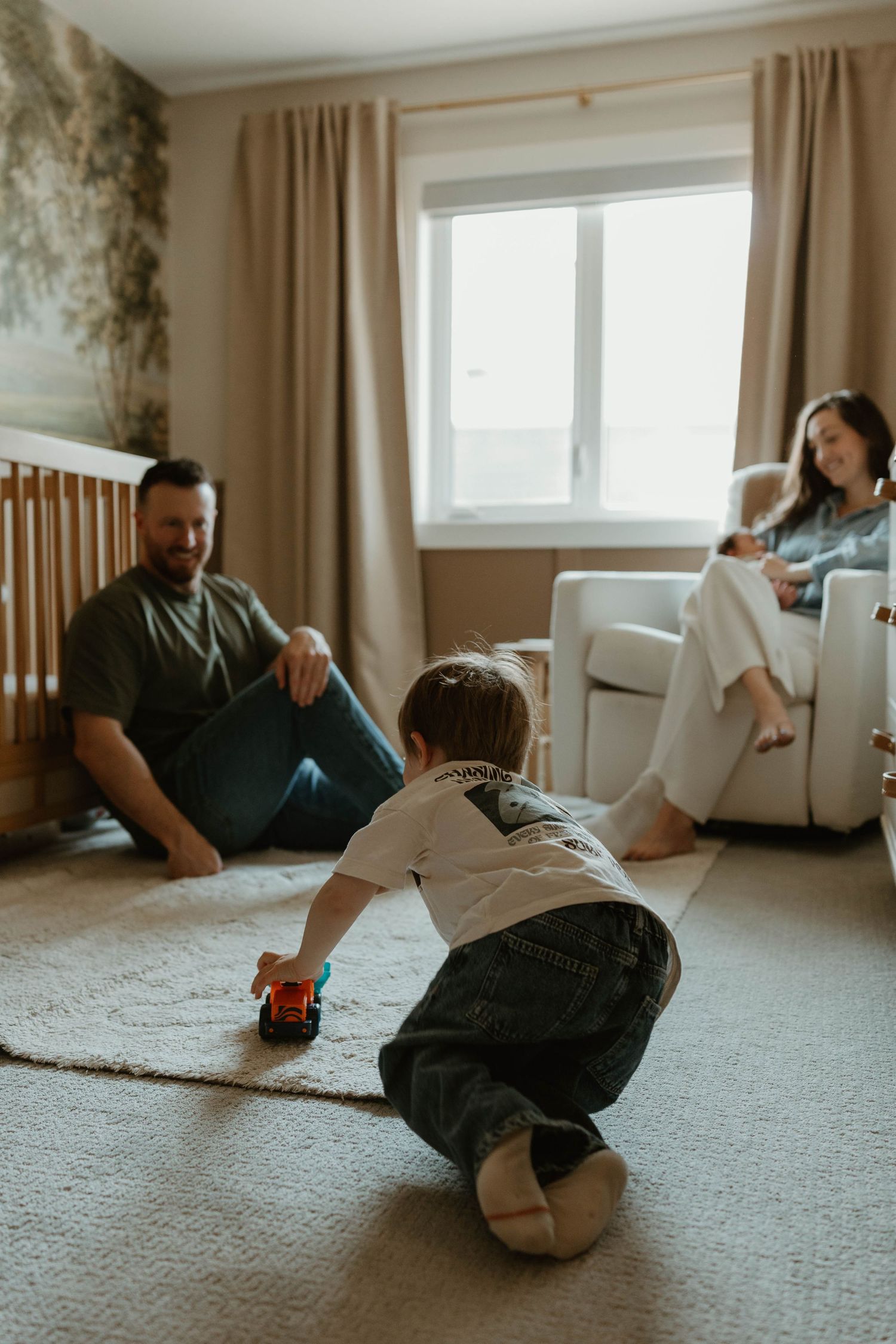 A toddler plays with a toy car on a carpet while parents relax and watch in a cozy neutral-toned living room.