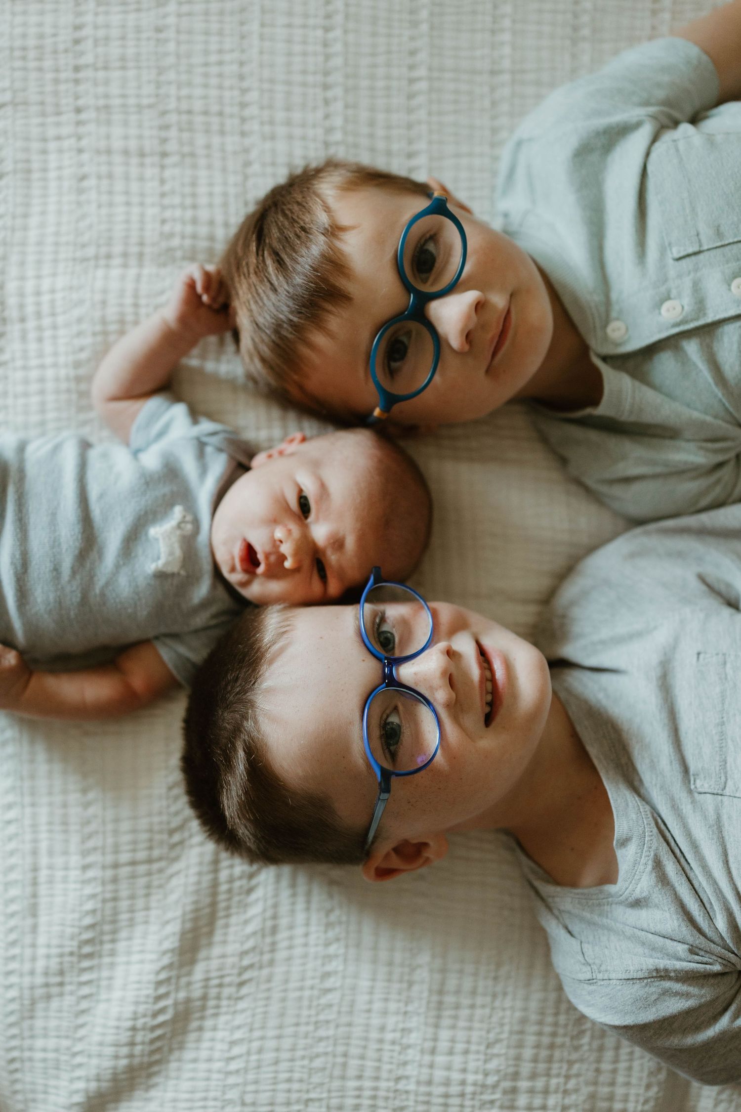 Three young boys lying together on a white bed, two wearing matching blue glasses, sharing a sweet sibling moment.